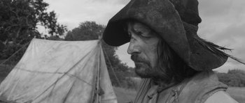 Movie still from “A Field in England” (2013), directed by Ben Wheatley – A person wearing a hat near a tent; Close Up shot, Over the shoulder angle