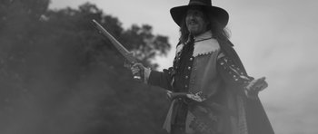 Movie still from “A Field in England” (2013), directed by Ben Wheatley – A man dressed as a musketeer holding a rifle; Medium shot, Low angle