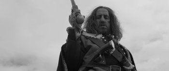 Movie still from “A Field in England” (2013), directed by Ben Wheatley – A man with long hair and a beard holding a gun; Medium shot, Low angle