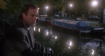 Movie still from “A Fish Called Wanda” (1988), directed by Charles Crichton – A man in a suit sitting in front of some boats; Medium shot, Low angle