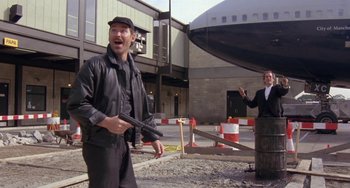 Movie still from “A Fish Called Wanda” (1988), directed by Charles Crichton – A man holding a gun in his hand while standing next to another man in front of a building; Wide shot, Over the shoulder angle