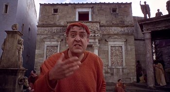 Movie still from “A Funny Thing Happened on the Way to the Forum” (1966), directed by Richard Lester – A man wearing a red hat and a red sweater is standing in front of an old building; Medium shot, Low angle