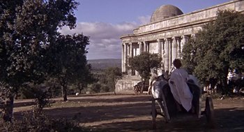 Movie still from “A Funny Thing Happened on the Way to the Forum” (1966), directed by Richard Lester – Two people are sitting on a bench in front of a building; Extreme Wide shot, Low angle