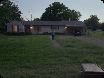 Movie still from “A Ghost Story” (2017), directed by David Lowery – A man standing in front of a house with a car parked in front of it; Extreme Wide shot, High angle