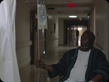 Movie still from “A Ghost Story” (2017), directed by David Lowery – A man sitting in a chair in a hospital hallway; Medium shot, High angle