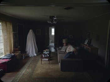 Movie still from “A Ghost Story” (2017), directed by David Lowery – A man sitting on a couch in a living room; Wide shot, Over the shoulder angle