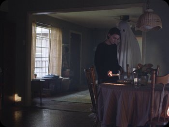 Movie still from “A Ghost Story” (2017), directed by David Lowery – A woman standing at a table in a room with candles; Medium shot, Over the shoulder angle