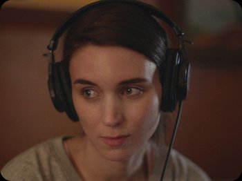 Movie still from “A Ghost Story” (2017), directed by David Lowery – A woman wearing headphones while sitting in a room; Close Up shot, Over the shoulder angle