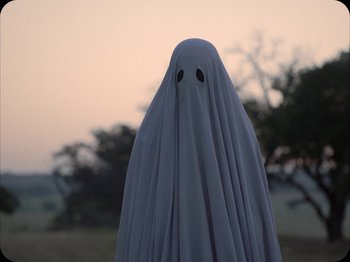 Movie still from “A Ghost Story” (2017), directed by David Lowery – A person in a ghost costume standing in a field; Medium shot, High angle