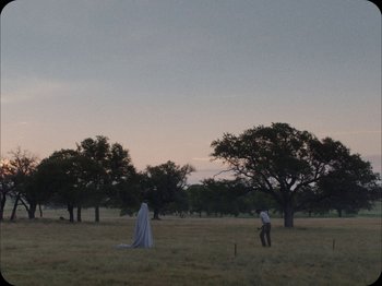 Movie still from “A Ghost Story” (2017), directed by David Lowery – Two people in a field with trees in the background; Extreme Wide shot, Low angle