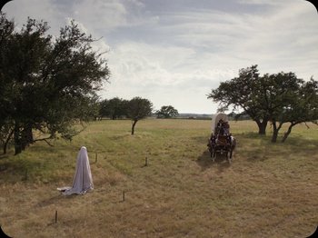 Movie still from “A Ghost Story” (2017), directed by David Lowery – A man in a white robe standing next to a horse drawn carriage; Extreme Wide shot, High angle