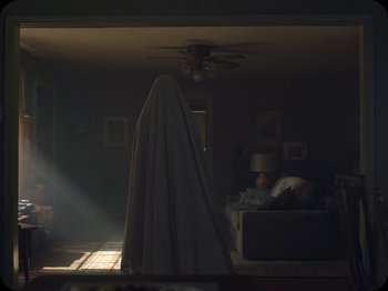 Movie still from “A Ghost Story” (2017), directed by David Lowery – A person in a living room wearing a ghost costume; Wide shot, Low angle
