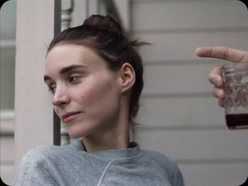 Movie still from “A Ghost Story” (2017), directed by David Lowery – A young woman is pointing to the side of her face; Close Up shot, Over the shoulder angle