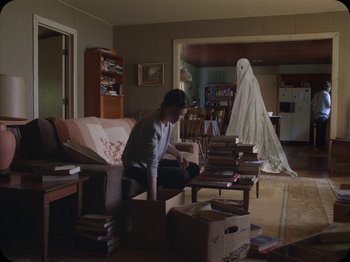 Movie still from “A Ghost Story” (2017), directed by David Lowery – A man sitting on top of a couch in a living room; Wide shot, Over the shoulder angle