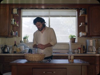 Movie still from “A Ghost Story” (2017), directed by David Lowery – A man standing in a kitchen preparing food; Medium shot, Low angle