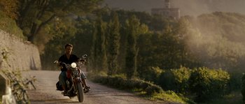 Movie still from “A Good Year” (2006), directed by Ridley Scott – A man riding a motorcycle down a dirt road near trees; Wide shot, Low angle