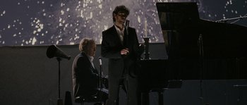 Movie still from “A Good Year” (2006), directed by Ridley Scott – A man standing at a piano with a microphone; Medium shot, Over the shoulder angle