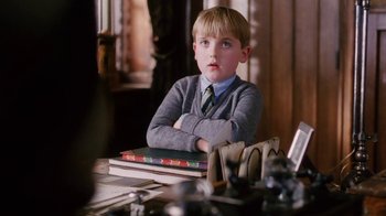 Movie still from “A Handful of Dust” (1988), directed by Charles Sturridge – A young boy sitting at a table with a stack of books; Medium shot, Over the shoulder angle