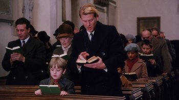 Movie still from “A Handful of Dust” (1988), directed by Charles Sturridge – A man in a black suit is holding a book; Medium shot, Low angle