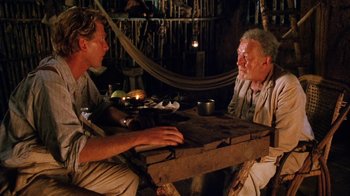 Movie still from “A Handful of Dust” (1988), directed by Charles Sturridge – Two men sitting at a wooden table in a hut; Medium shot, Over the shoulder angle