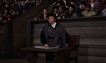 Movie still from “A Man for All Seasons” (1966), directed by Fred Zinnemann – A man sitting at a table in front of a group of people; Medium shot, High angle