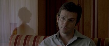 Movie still from “A Mighty Heart” (2007), directed by Michael Winterbottom – A person sitting on a couch wearing glasses; Close Up shot, Over the shoulder angle