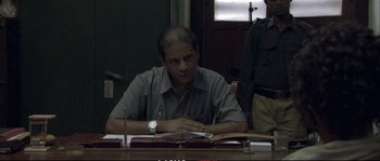 Movie still from “A Mighty Heart” (2007), directed by Michael Winterbottom – A man sitting at a table with papers in front of him; Medium shot, Over the shoulder angle