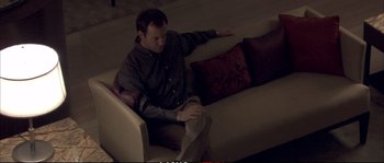 Movie still from “A Mighty Heart” (2007), directed by Michael Winterbottom – A man sitting on top of a white couch next to pillows; Medium shot, High angle