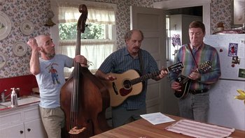 Movie still from “A Mighty Wind” (2003), directed by Christopher Guest – A group of people standing around a table with a guitar; Wide shot, Low angle