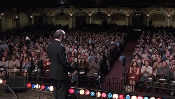 Movie still from “A Mighty Wind” (2003), directed by Christopher Guest – A man standing on a stage in front of an audience; Wide shot, High angle