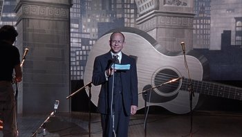 Movie still from “A Mighty Wind” (2003), directed by Christopher Guest – A man standing in front of a microphone in front of a guitar; Medium shot, High angle