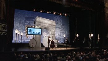 Movie still from “A Mighty Wind” (2003), directed by Christopher Guest – A stage with a man on a stage with a microphone and a cello; Extreme Wide shot, Low angle