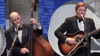 Movie still from “A Mighty Wind” (2003), directed by Christopher Guest – A man playing a guitar while a man plays a double bass; Medium shot, Low angle