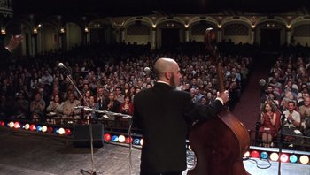 Movie still from “A Mighty Wind” (2003), directed by Christopher Guest – A man with a beard playing a contrabass in front of an audience; Wide shot, Low angle
