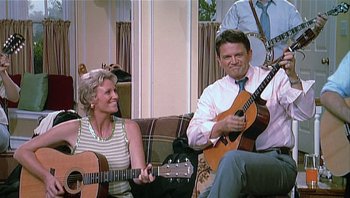 Movie still from “A Mighty Wind” (2003), directed by Christopher Guest – A man and a woman sitting on a couch with guitars; Medium shot, High angle