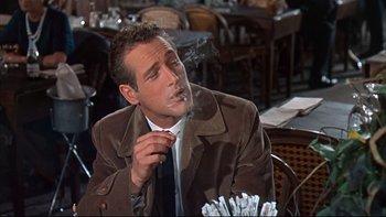 Movie still from “A New Kind of Love” (1963), directed by Melville Shavelson – A man smoking a cigarette while sitting at a table; Medium shot, Over the shoulder angle