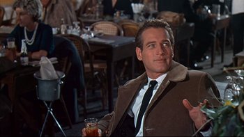 Movie still from “A New Kind of Love” (1963), directed by Melville Shavelson – A man sitting in front of a table holding a drink; Medium shot, Over the shoulder angle