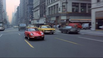 Movie still from “A New Leaf” (1971), directed by Elaine May – A red sports car driving down a street past tall buildings; Wide shot, High angle