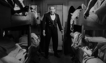 Movie still from “A Night to Remember” (1958), directed by Roy Ward Baker – A black and white photo of a man in a suit standing in front of a bunch of bunkbeds; Medium shot, High angle