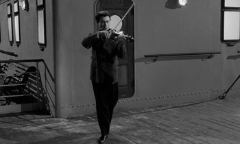 Movie still from “A Night to Remember” (1958), directed by Roy Ward Baker – A black and white photo of a man playing the violin; Wide shot, Low angle