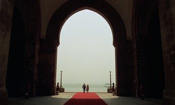 Movie still from “A Passage to India” (1984), directed by David Lean – Two people are standing on a red carpet in front of the water; Extreme Wide shot, Low angle