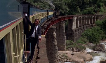 Movie still from “A Passage to India” (1984), directed by David Lean – A man standing on the side of a train on a bridge; Wide shot, Low angle