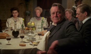 Movie still from “A Room with a View” (1985), directed by James Ivory – An older man sitting at a dinner table with other people; Medium shot, Over the shoulder angle