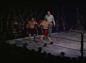 Movie still from “A Star Is Born” (1937), directed by Victor Fleming – Two men in red and blue shorts fighting in a boxing ring; Wide shot, High angle