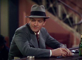 Movie still from “A Star Is Born” (1937), directed by Victor Fleming – A man wearing a hat and a suit sitting at a table with a drink; Close Up shot, High angle