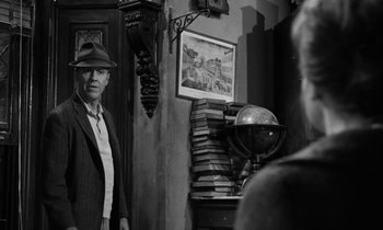 Movie still from “A Thousand Clowns” (1965), directed by Fred Coe – A man in a suit and hat standing in front of a stack of books; Medium shot, Over the shoulder angle