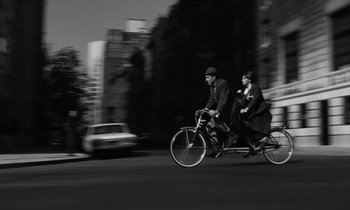 Movie still from “A Thousand Clowns” (1965), directed by Fred Coe – Two people riding bicycles down a street with a car in the background; Wide shot, High angle