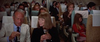 Movie still from “A Touch of Class” (1973), directed by Melvin Frank – A woman holding a glass of wine in a crowded airplane; Medium shot, Over the shoulder angle