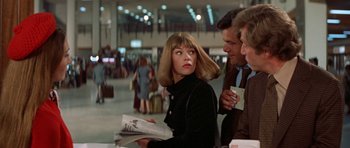 Movie still from “A Touch of Class” (1973), directed by Melvin Frank – A woman is holding a newspaper while standing next to two other people; Medium shot, Over the shoulder angle
