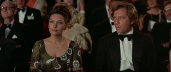 Movie still from “A Touch of Class” (1973), directed by Melvin Frank – A man and a woman standing next to each other in front of a crowd; Medium shot, Low angle